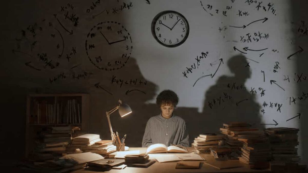 A student sitting at a desk late at night surrounded by tall stacks of books and clock shadows on the wall, illustrating academic pressure and burnout.