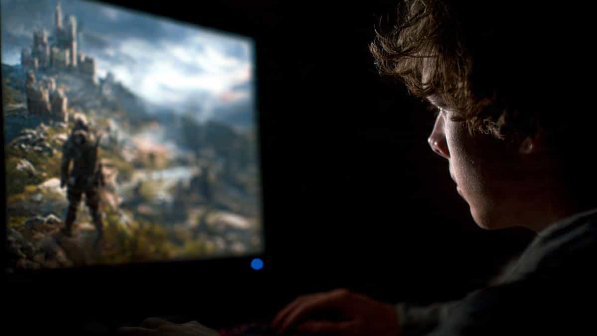 A calm person sits in a dark room looking at a game screen showing a fantasy RPG character standing on a hill overlooking a distant castle. The screen glow softly lights their face. Represents how RPG games let players become someone else and explore identity.