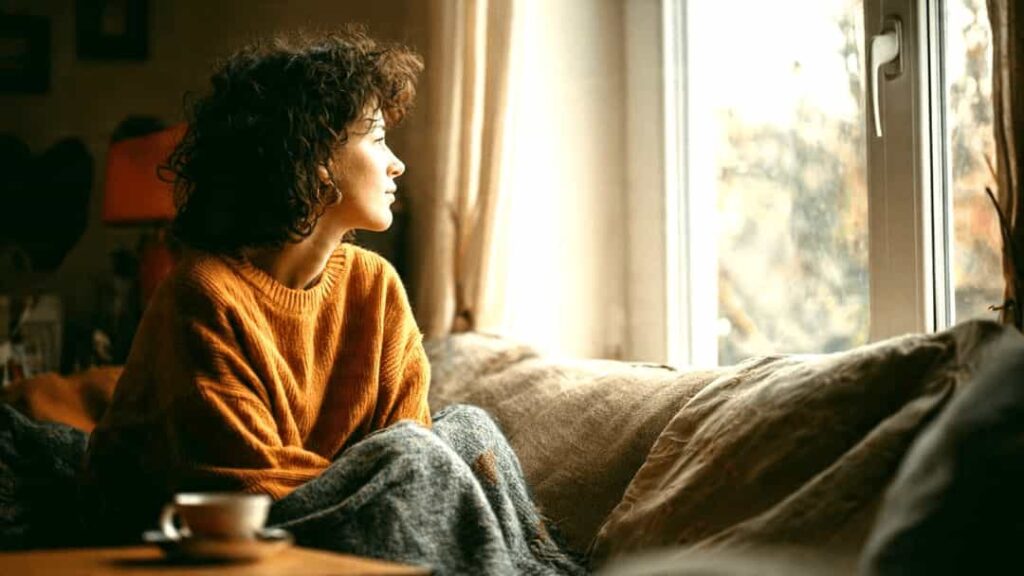 Person sitting alone on a sofa looking out a window, choosing peaceful distance from a difficult friendship
